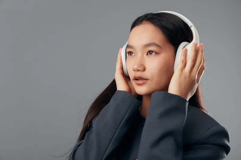 Young woman wearing white headphones listening to music or podcast focused on sound with closed lips isolated on gray background in casual dark jacket for leisure or entertainment.