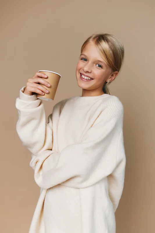 Smiling girl wearing a cozy white sweater holds a paper coffee cup, standing against a neutral beige backdrop, capturing a warm, inviting studio portrait.
