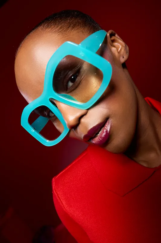 Close up portrait of a black woman with dark skin wearing oversized turquoise glasses and a red jacket. Studio lighting emphasizes fashion and confident mood on red background.