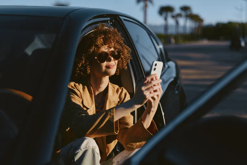 A woman with curly hair sits in a car, taking a casual selfie with a smartphone as bright sun lights the scene, conveying a relaxed lifestyle moment with retro film color.