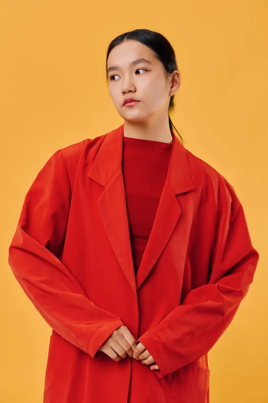 Young woman wearing oversized red blazer and matching sweater stands against vibrant orange background, looking sideways with serious expression and dark hair tied back neatly.