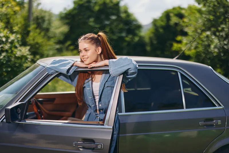 Young woman enjoying a sunny day leaning out of a vintage car, wearing a denim jacket, with a cheerful expression in a green outdoor setting.