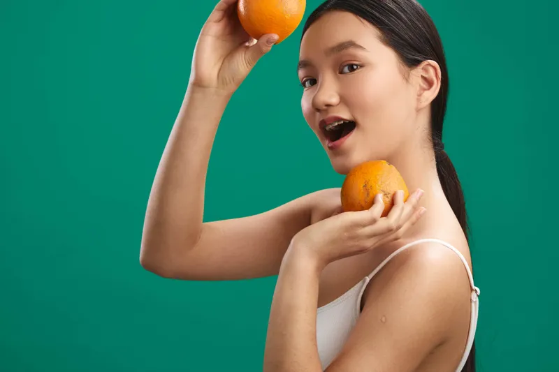 An Asian woman poses with oranges in a bright studio, smiling playfully as she experiments with fruit props, creating a fresh, vibrant lifestyle image.