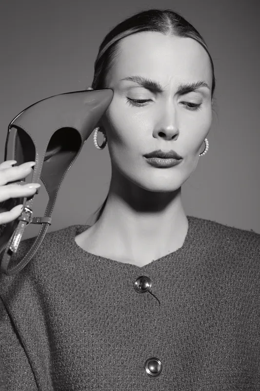 Caucasian woman with fair skin and slicked hair holds high heel shoe near her head with closed eyes. Monochrome portrait in studio lighting. Concept of fashion, style, and emotion.