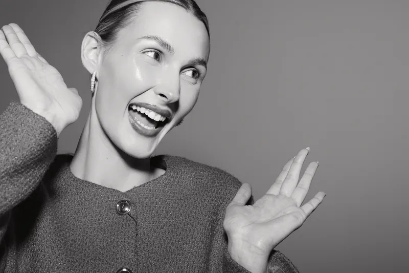 Smiling white woman with light skin and short hair poses happily with raised hands on grey background. Portrait of cheerful young female expressing joy and excitement in studio lighting.