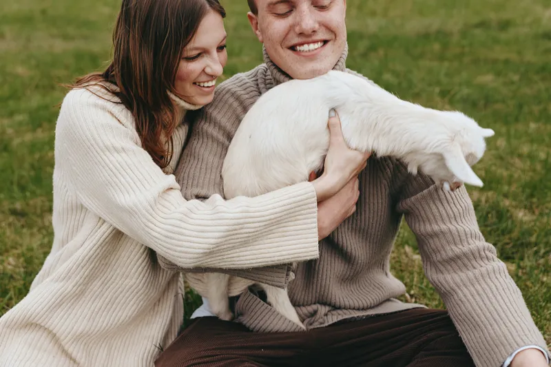 Couple smiling while holding a white goat kid in a grassy meadow. Warm natural light highlights a candid rural lifestyle moment, celebrating animal bond and gentle affection.
