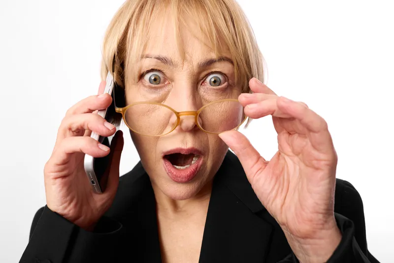 Woman surprised on phone, holding eyeglasses with both hands, wide open mouth, studio lighting, white background, communication moment, shocked expression