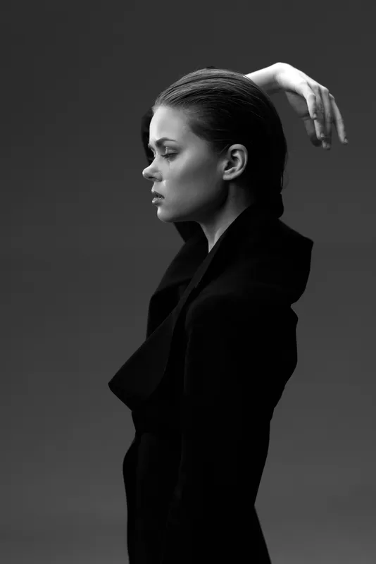Black and white profile of a stylish woman with braided hair wearing a dark coat, expressing calm and confidence in a minimalist studio setting.