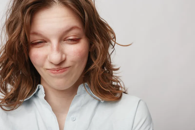 Young woman with wavy hair makes funny face with closed eyes and slight smile, wearing white shirt on plain background. Concept of humor, emotion, and casual portrait.