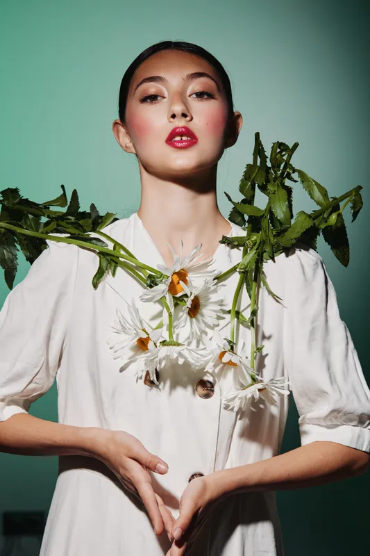 Young woman with vibrant glossy lipstick wearing white dress adorned with fresh daisies. Studio portrait on green background. Concept for beauty, creative, and fashion editorial.