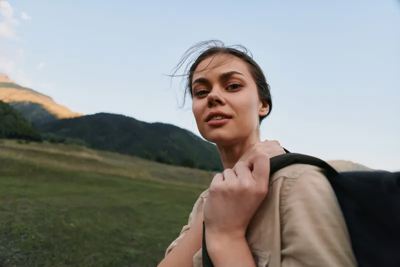 Young woman with dark hair standing outdoors in a natural landscape with mountains and grass. Casual lifestyle concept with soft natural lighting and calm mood.