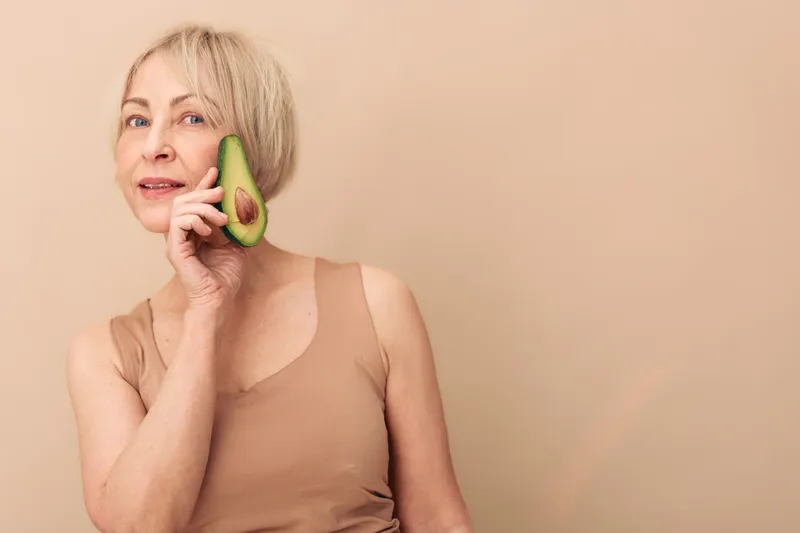 Mature woman with short blonde hair holds avocado half near face, smiling gently. Neutral beige background and soft lighting create natural skincare and healthy lifestyle concept.