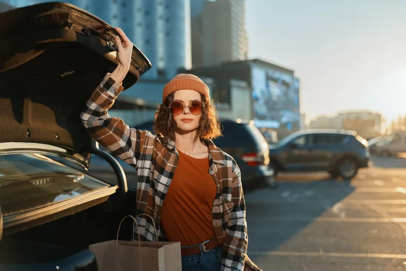 woman shopping by car in parking city at sunset, casual style with plaid shirt, beanie and sunglasses by trunk. Urban portrait capturing authenticity, candid lifestyle and golden hour glow