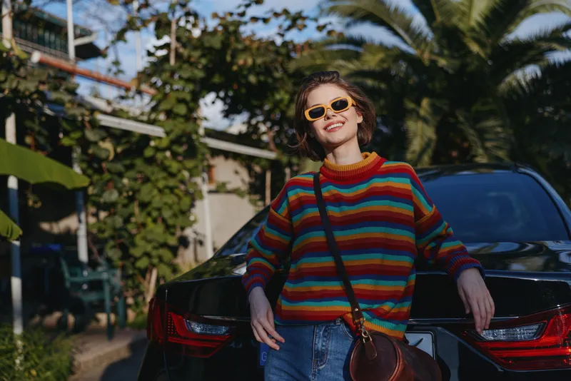 Woman in a colorful striped sweater and yellow sunglasses leaning against a black car, smiling confidently, wearing denim jeans and crossbody bag in a tropical outdoor urban street setting.