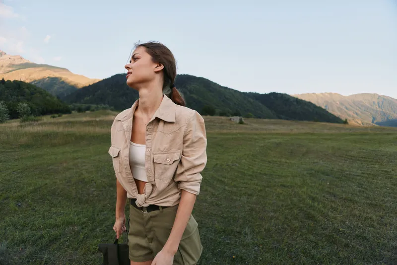 A young woman stands in a wide green field with distant mountains, wearing a beige shirt and olive shorts, looking upward with a relaxed, confident mood as the sun warms the landscape.