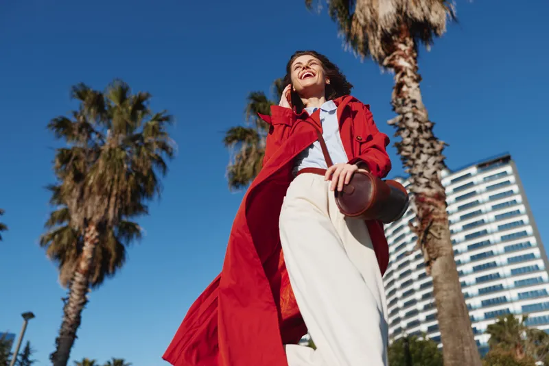 A stylish young woman in a red coat and white pants stands outdoors under a bright blue sky, palm trees framing a modern backdrop and a confident, spirited mood.