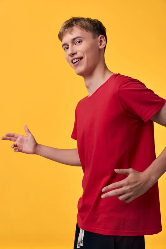 Young man in a red t shirt stands against a vibrant yellow backdrop, presenting a dynamic pose with open hands and a bright smile, conveying energy, confidence, and casual style.