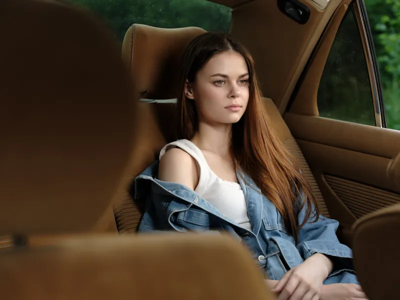 Young woman with long hair in denim jacket sitting in a car, reflecting on life, exuding calmness and contemplation in a natural green background.