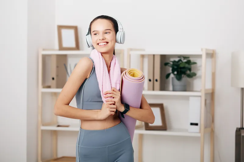 Woman fitness enthusiast in a home gym, wearing activewear and headphones, holding a rolled yoga mat and towel, ready for a workout, bright calm room, energy and motivation.