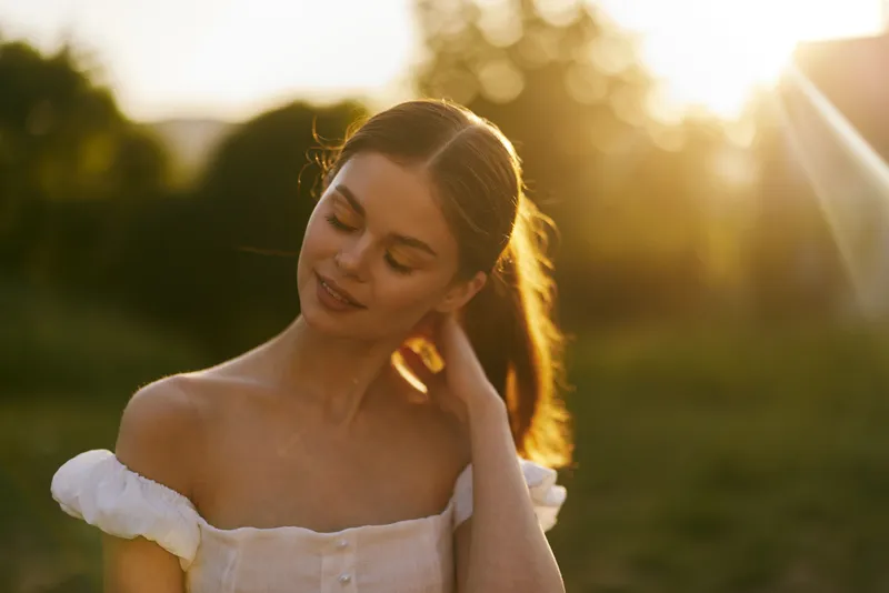 Beautiful young woman with long hair enjoying a sunny evening in a white dress, displaying serenity and warmth amidst a natural backdrop