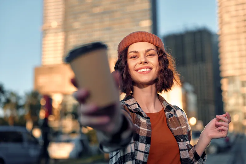 Woman holding coffee cup and smiling in urban street, candid authenticity and lifestyle portrait at golden hour, golden hour glow, mindful living and emotional storytelling mood.