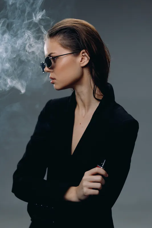 Stylish woman with sunglasses vaping smoke in a dark setting, wearing black, confident and modern look.