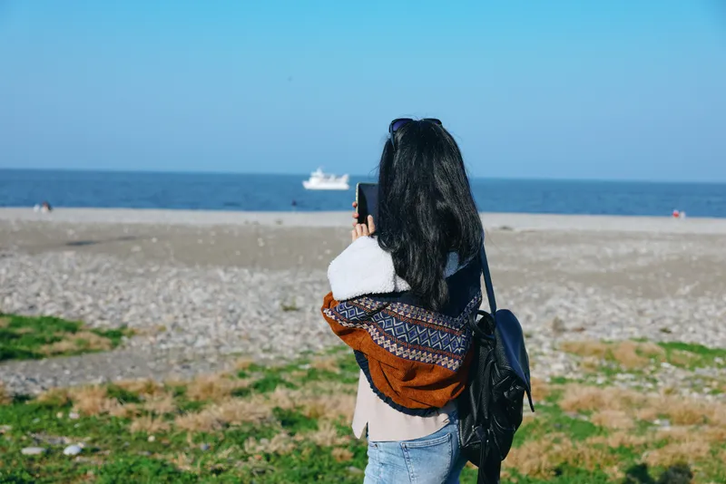Individual with backpack standing on a sandy beach, capturing the sea with a smartphone, blue horizon over calm water, casual attire, travel mood, outdoor photography in bright daylight
