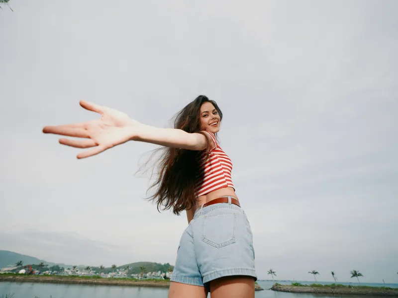 A smiling woman in a striped crop top and denim shorts extends her arms toward the camera, capturing bright, carefree summer energy against a seaside sky.