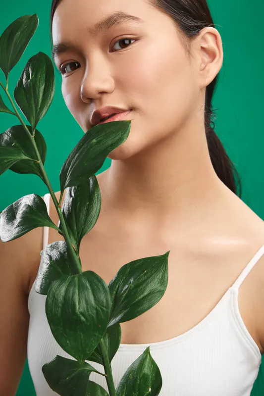 Asian woman holds a leafy plant against a vivid green backdrop, portrait shot with soft lighting. Her calm expression and graceful pose create a fresh, natural mood with a modern, minimal vibe.