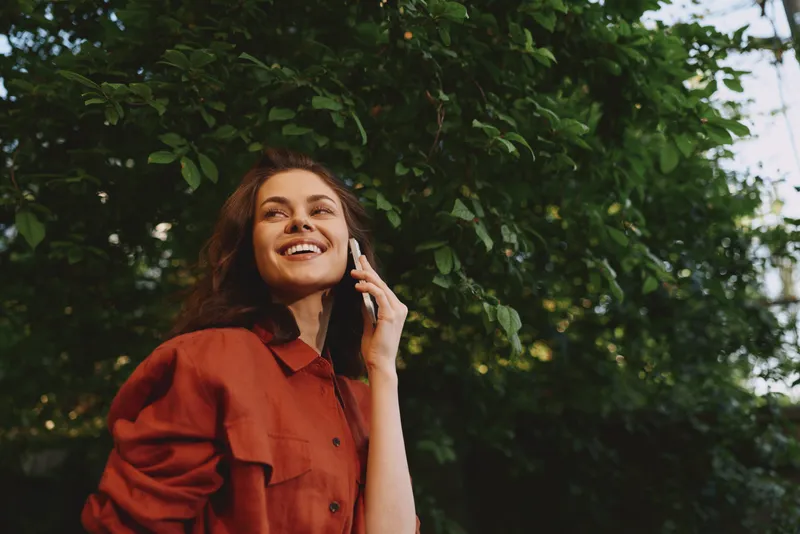 Bright woman outdoors in a red shirt talks on her phone, smiling, capturing a joyful lifestyle moment in a natural green background, casual candid portrait shot