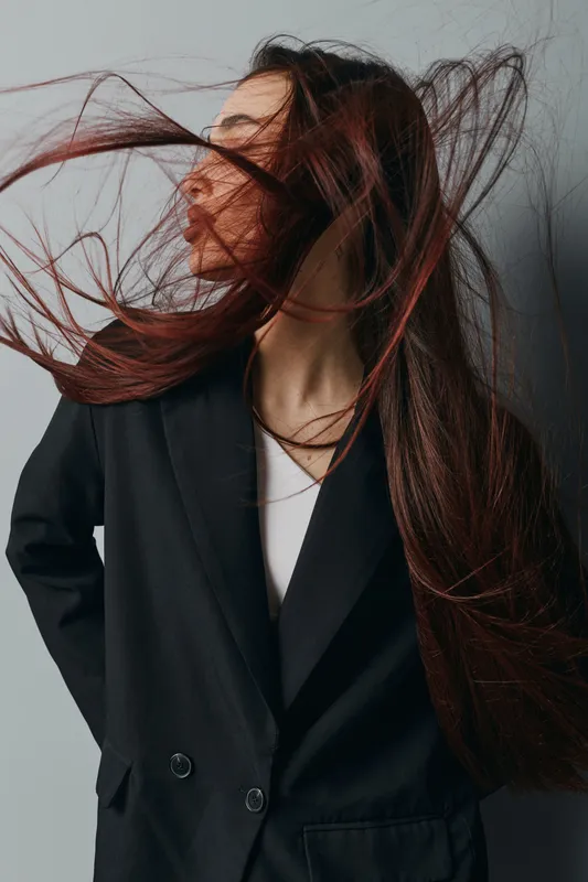 Woman with long hair blowing in wind wearing black blazer and white shirt, posing in studio with grey background. Fashion portrait with dynamic movement and natural expression.