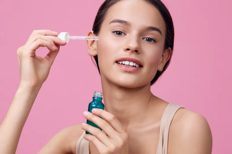 Young woman applying facial serum with dropper on a pink background, showcasing skin care and beauty concepts with a fresh, healthy glow