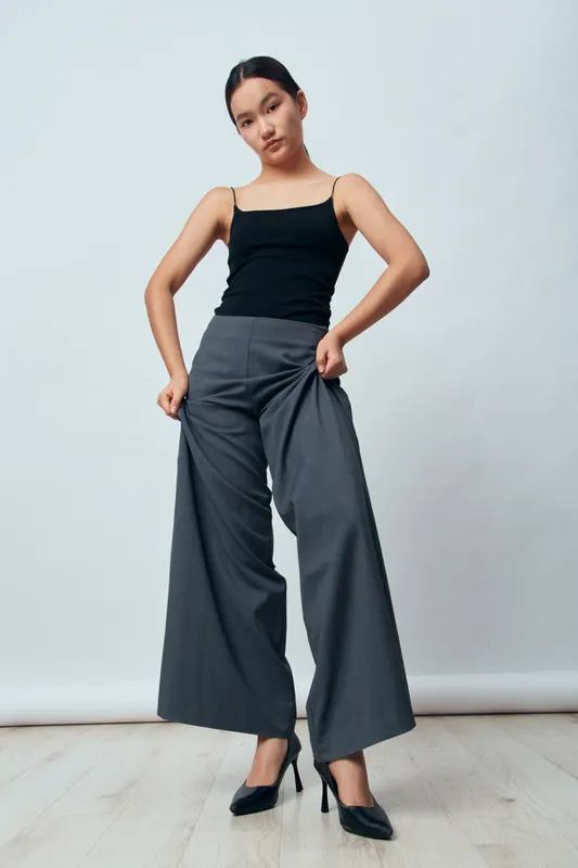 Young woman standing indoors wearing wide pants and black tank top with heels on light wooden floor and neutral background for fashion style. Female model posing with confident expression.