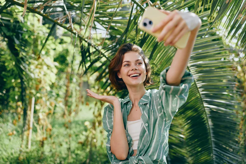 Bright portrait of a smiling woman taking a selfie in a sunny tropical garden, wearing a green shirt, surrounded by lush plants and vibrant foliage.