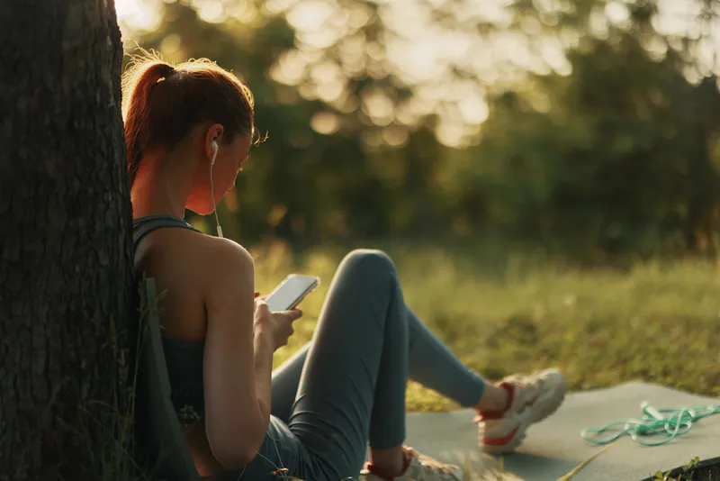 Young woman in sportswear enjoying music while sitting on a yoga mat outdoors, blending relaxation and fitness in a serene natural setting