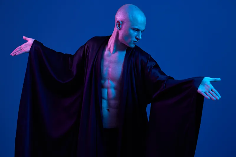 Artistic man in black robe with dramatic pose, showcasing strength and elegance under vibrant blue light. A captivating contrast between shadows and colors enhances the mood.