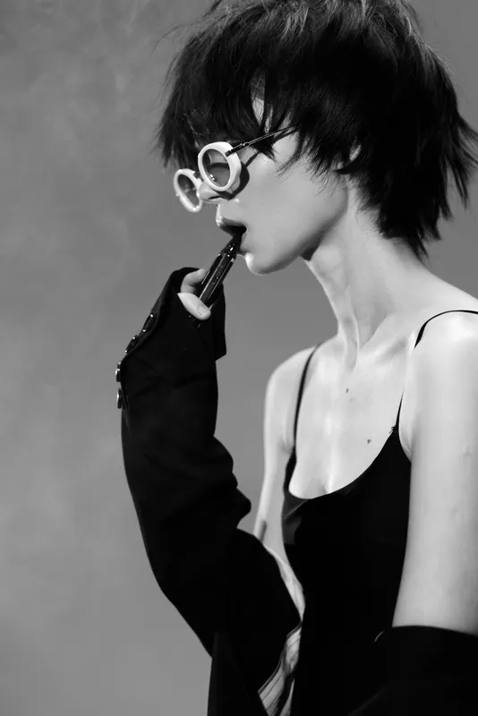 Black and white portrait of a stylish woman wearing round glasses and gloves, posing thoughtfully against a plain background.