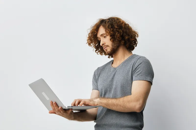 Young man with curly hair using a laptop, wearing a gray T shirt in a minimalistic room, conveying focus and concentration with a modern, neutral background