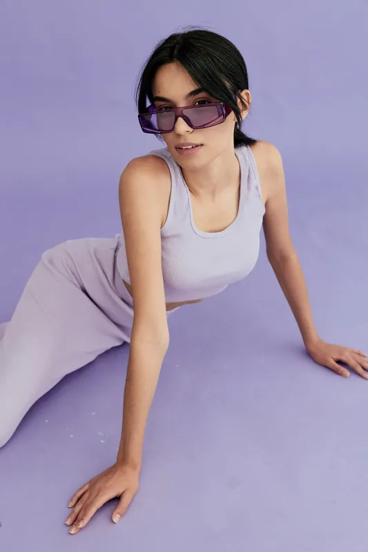 young woman wearing purple sunglasses and lavender outfit poses on lilac floor with soft lighting, presenting modern fashion and stylish summer look