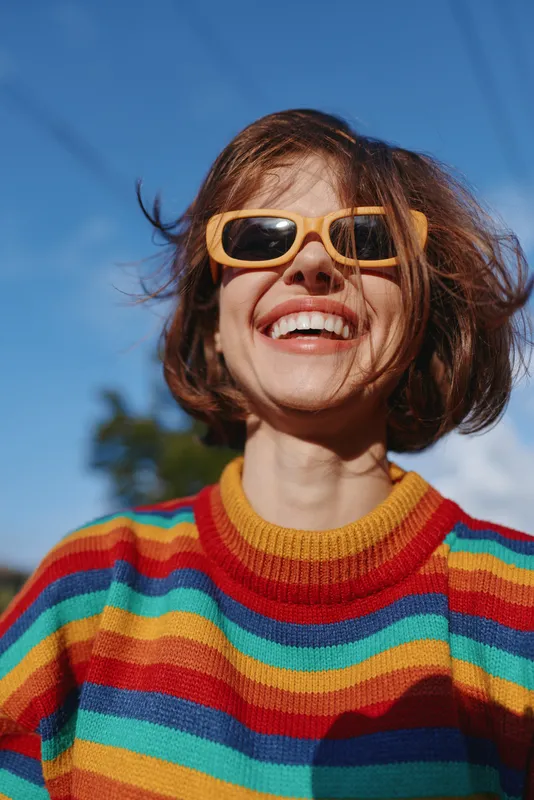 Woman smiling in rainbow sweater and yellow sunglasses, late 20s brunette with short hair, outdoor portrait under blue sky, joyful lifestyle fashion travel vibe.