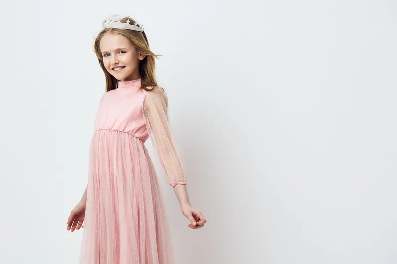 Smiling girl in a pink dress with sheer sleeves and a crown, exuding joy and elegance in a light, neutral background for children s fashion photography