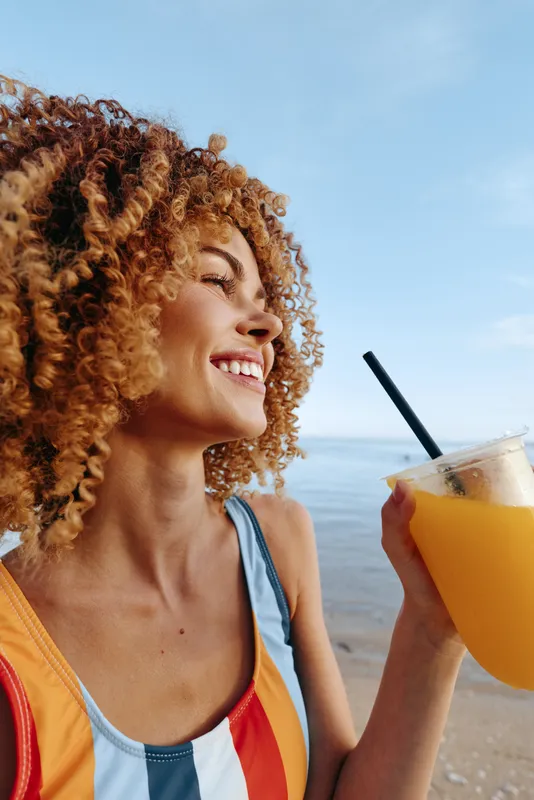 Smiling woman with curly hair holding a plastic cup with orange juice on a beach. Sunny day with clear blue sky, casual summer outfit, and relaxed mood outdoors.