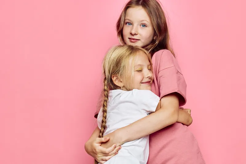 Two young girls embracing against a pink background. The older girl looks at the camera while the younger one closes her eyes smiling. Concept for love, siblings, family.