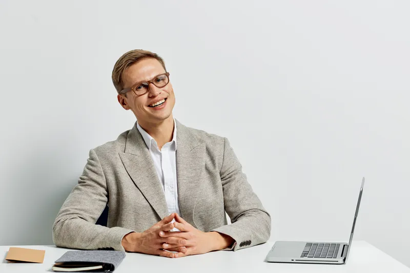 Smiling young man with glasses sitting at a desk with a laptop and notepad, wearing a gray blazer and white shirt, isolated on plain white background. Business, corporate, professional, technology, ed