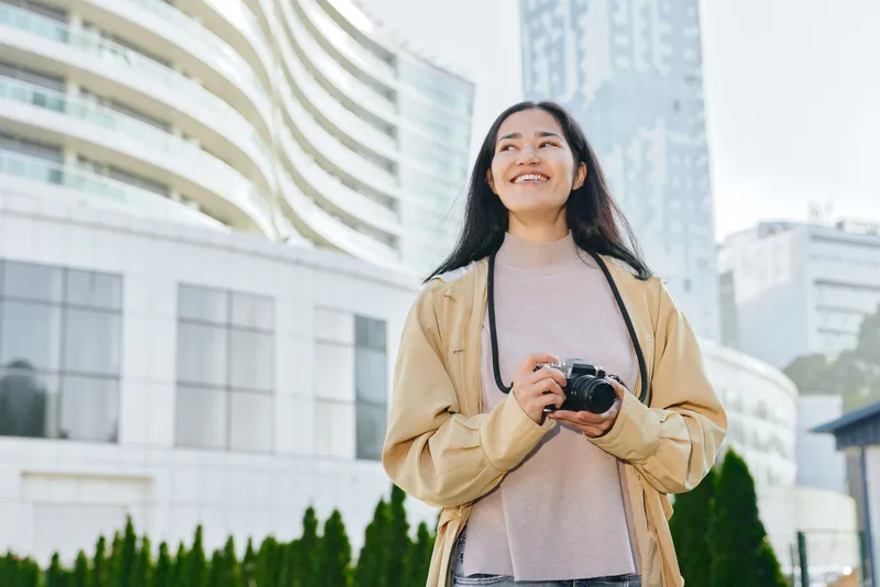 Smiling woman holding a camera outdoors in an urban cityscape, buildings behind her, jacket and sweater, conveying travel, photography, and exploration.