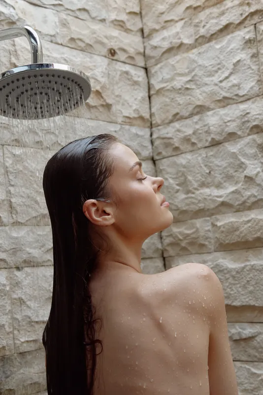 young woman under outdoor shower with wet hair, relaxed posture, natural stone wall background, serene moment, warm lighting