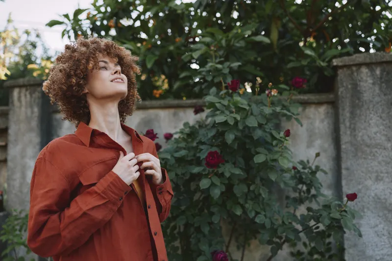Portrait of a woman with curly hair wearing a red jacket in a garden with blooming roses, conveying confidence, nature, and relaxed lifestyle through soft outdoor light