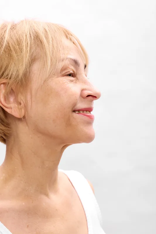 Woman closeup profile smile, blonde senior portrait, soft daylight, calm happy expression, gentle skin, skincare wellness moment, bright minimal background