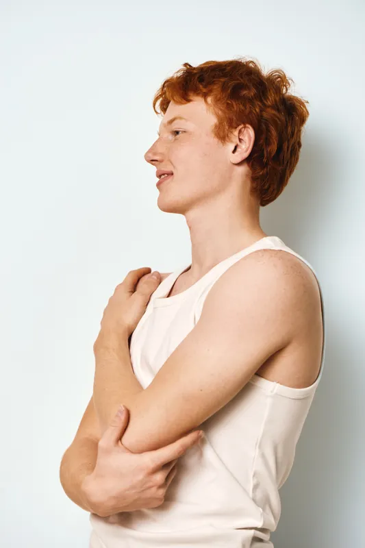 Young man with red hair and freckles wearing white tank top standing in profile against plain background with relaxed expression and folded arms.