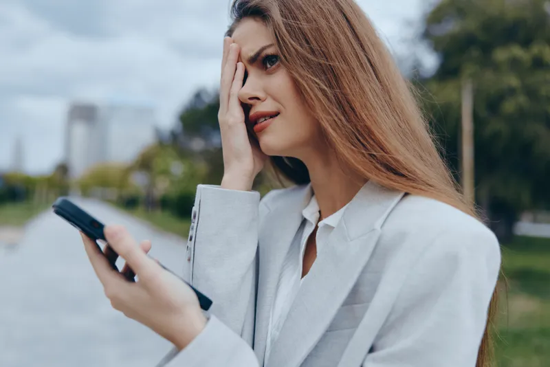 Woman in white blazer experiencing distress with smartphone outdoors in urban park conveying financial crisis emotions and stress